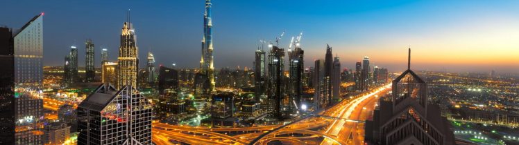 Panorama Dubaju o zmierzchu z widokiem na Burj Khalifa, nowoczesne wieżowce i wielopoziomowe skrzyżowania autostrad rozświetlone nocnymi światłami.
