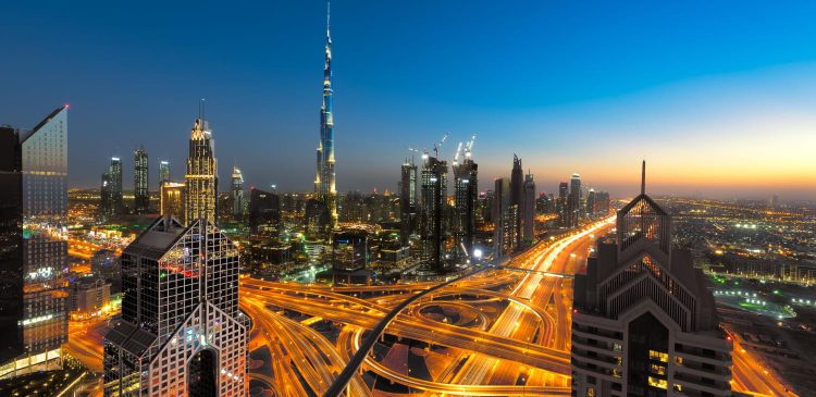 Panorama Dubaju o zmierzchu z widokiem na Burj Khalifa, nowoczesne wieżowce i wielopoziomowe skrzyżowania autostrad rozświetlone nocnymi światłami.