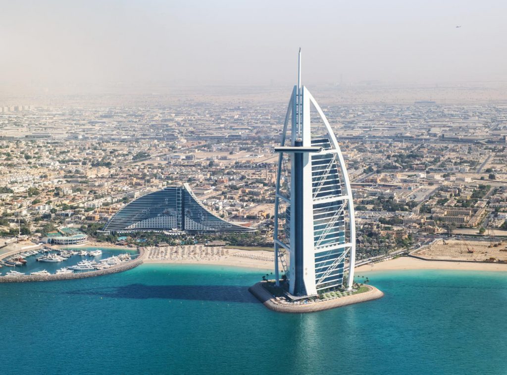 Widok z lotu ptaka na Burj Al Arab w Dubaju, luksusowy hotel na sztucznej wyspie, turkusowe morze, plażę i nowoczesną zabudowę miasta w tle.