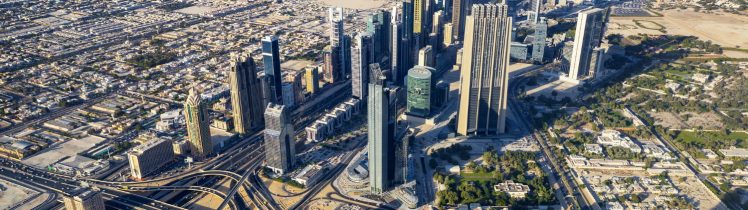 Panorama Dubaju z lotu ptaka przedstawiająca gęstą zabudowę wieżowców, rozbudowany węzeł drogowy oraz Zatokę Perską w tle.