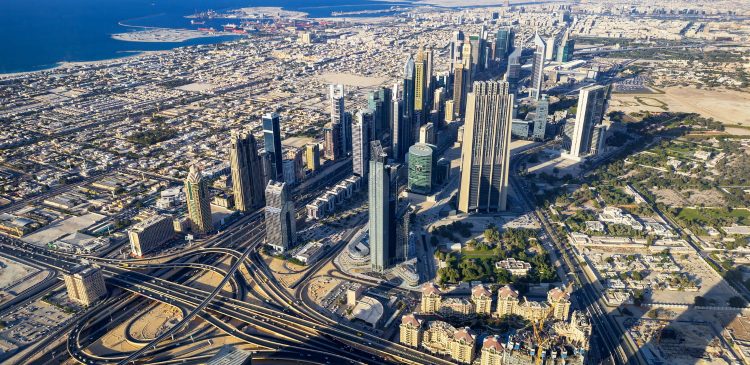 Panorama Dubaju z lotu ptaka przedstawiająca gęstą zabudowę wieżowców, rozbudowany węzeł drogowy oraz Zatokę Perską w tle.