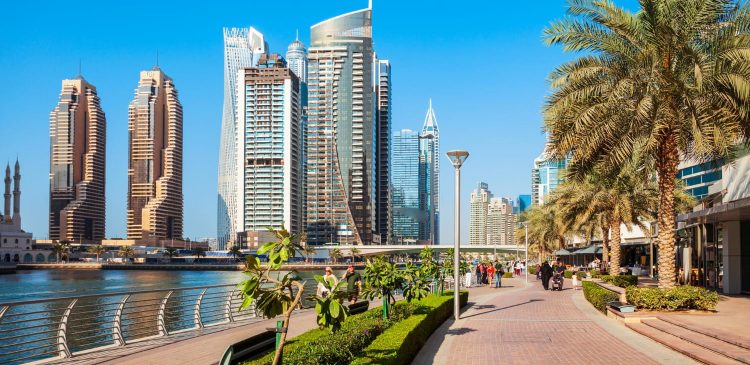Promenada nad wodą w Dubaju z palmami, spacerującymi ludźmi i nowoczesnymi wieżowcami w tle w słoneczny dzień.