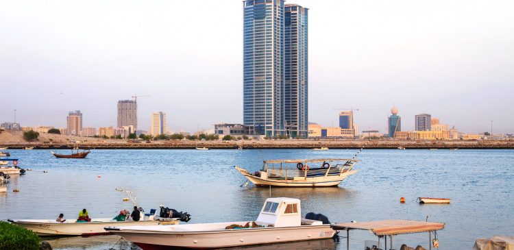 Nadbrzeże w Ras Al Khaimah z łodziami rybackimi na spokojnej wodzie i nowoczesnym wieżowcem w tle.