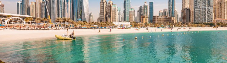 Plaża w Dubaju z turkusową wodą, jachtami i widokiem na nowoczesne wieżowce Dubai Marina w tle.