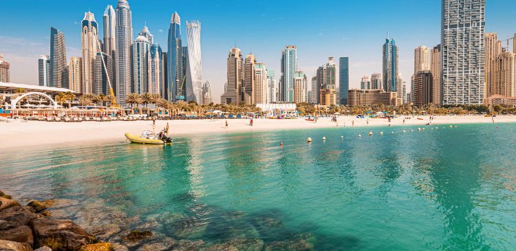Plaża w Dubaju z turkusową wodą, jachtami i widokiem na nowoczesne wieżowce Dubai Marina w tle.