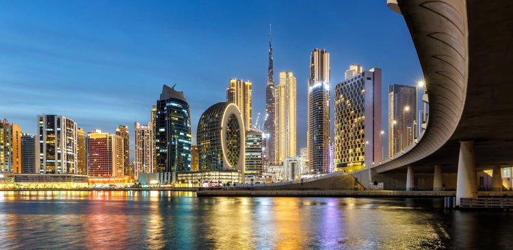 Nocna panorama Dubaju z oświetlonymi wieżowcami i Burj Khalifa w tle, odbijającymi się w wodzie kanału oraz estakadą po prawej stronie.