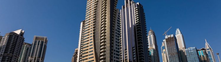 Nowoczesne wieżowce mieszkalne w Dubaju widziane z poziomu ulicy, z estakadą drogową na pierwszym planie i bezchmurnym niebem w tle.