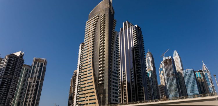 Nowoczesne wieżowce mieszkalne w Dubaju widziane z poziomu ulicy, z estakadą drogową na pierwszym planie i bezchmurnym niebem w tle.