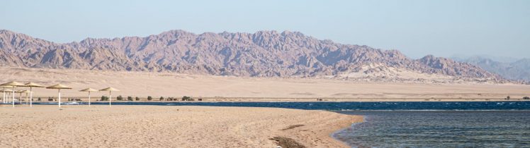 Piaszczysta plaża w Ras Al Khaimah z widokiem na spokojne morze i góry w tle, pustynny krajobraz z parasolami przeciwsłonecznymi