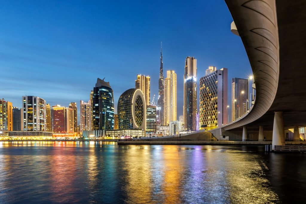 Wieczorna panorama Dubaju z oświetlonymi wieżowcami nad wodą i widokiem na Burj Khalifa w tle.