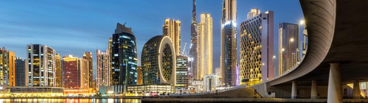Wieczorna panorama Dubaju z oświetlonymi wieżowcami nad wodą i widokiem na Burj Khalifa w tle.